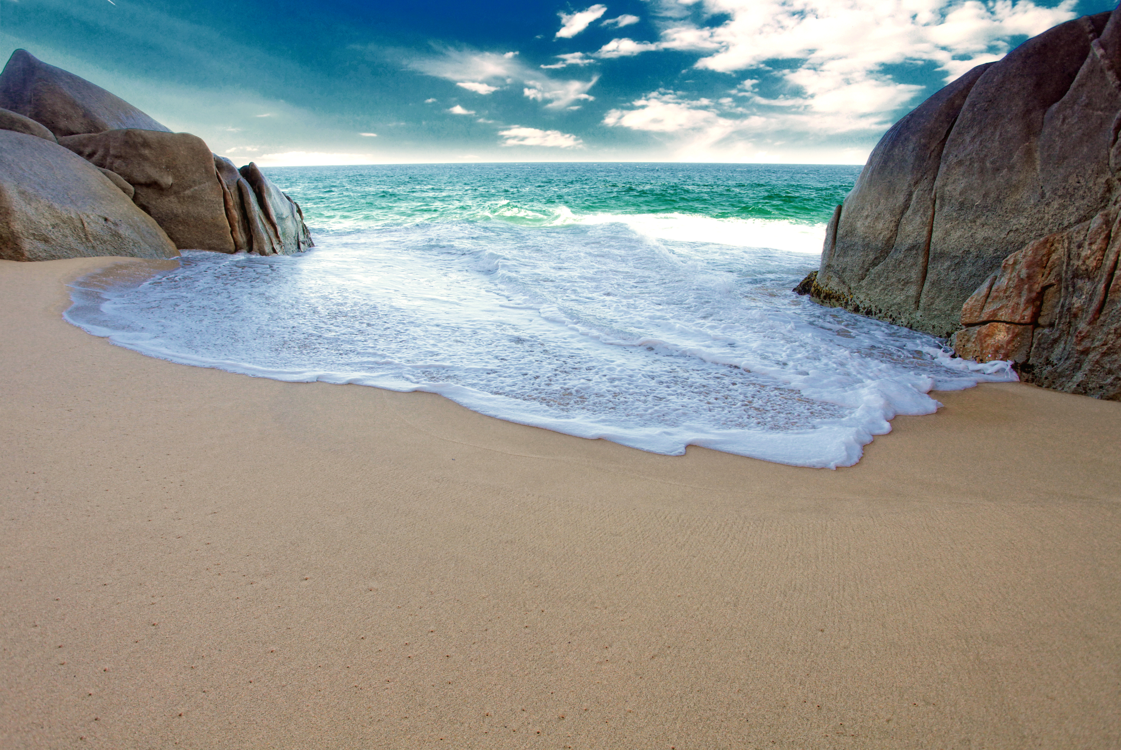 A scenic sandy beach featuring turquoise sea water and large grey rocks under a cloudy sky
