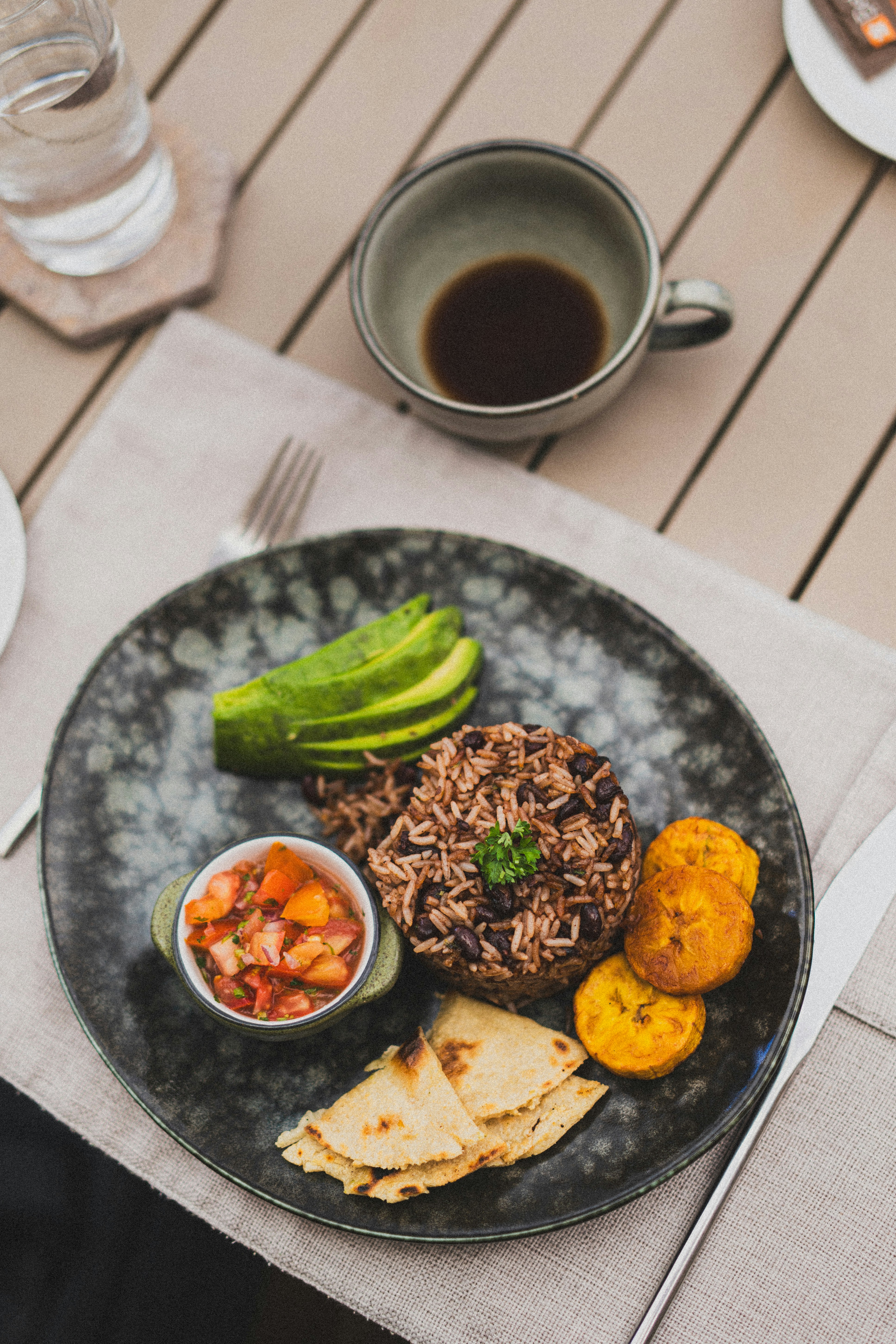 A grey plate holds rice and beans, sliced avocado, salsa, flatbread, and fried plantain, with coffee and water nearby.