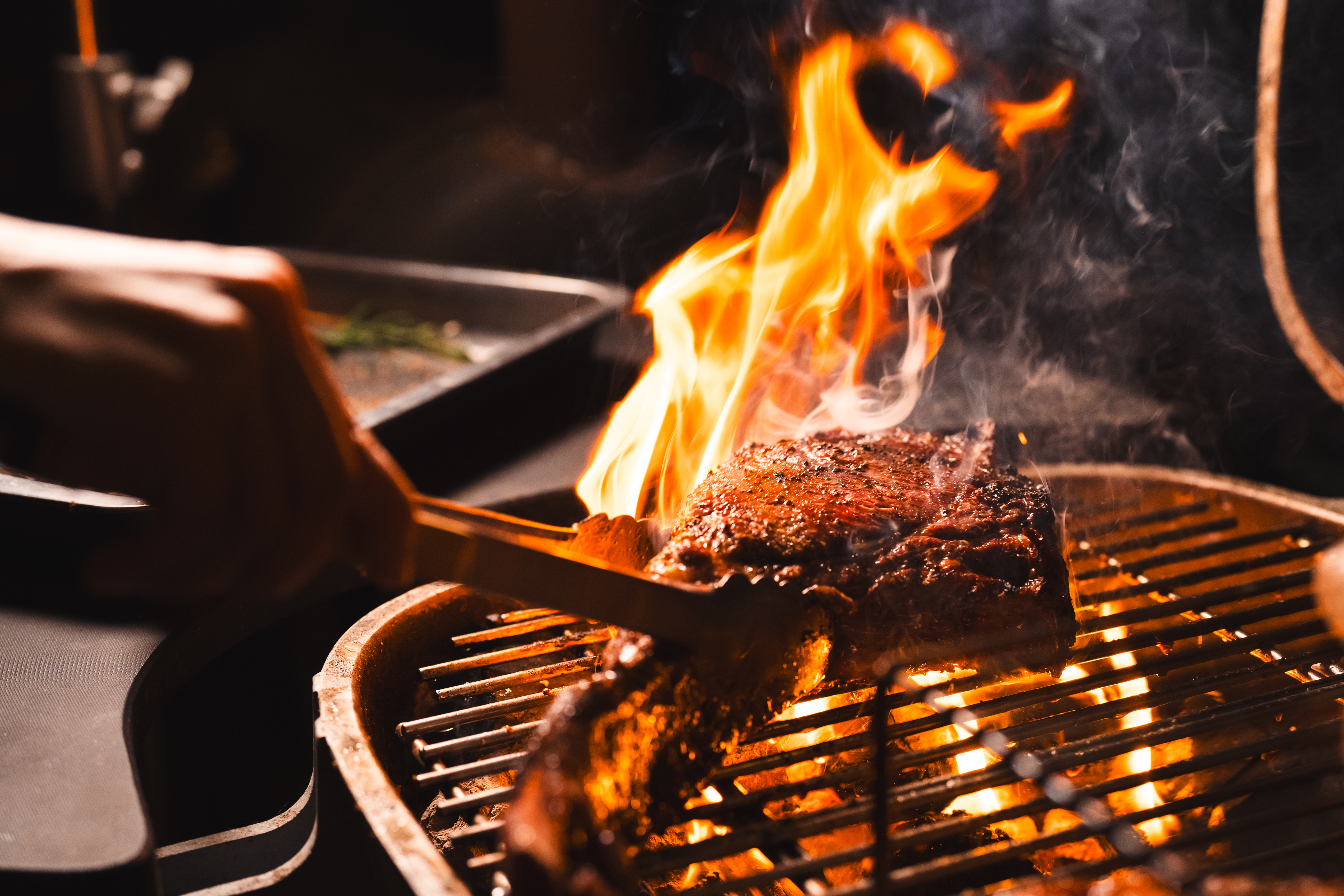 A thick steak cooks on a metal barbecue grill surrounded by bright orange flames and grey smoke