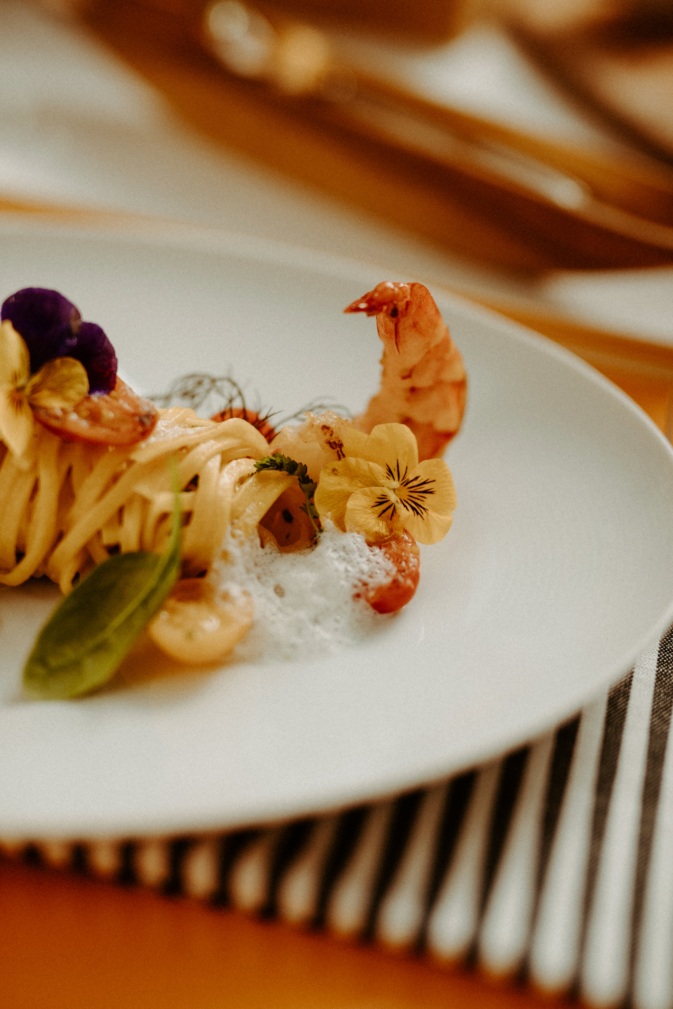 A gourmet pasta dish garnished with a prawn, edible flowers, and fresh herbs on a white plate.