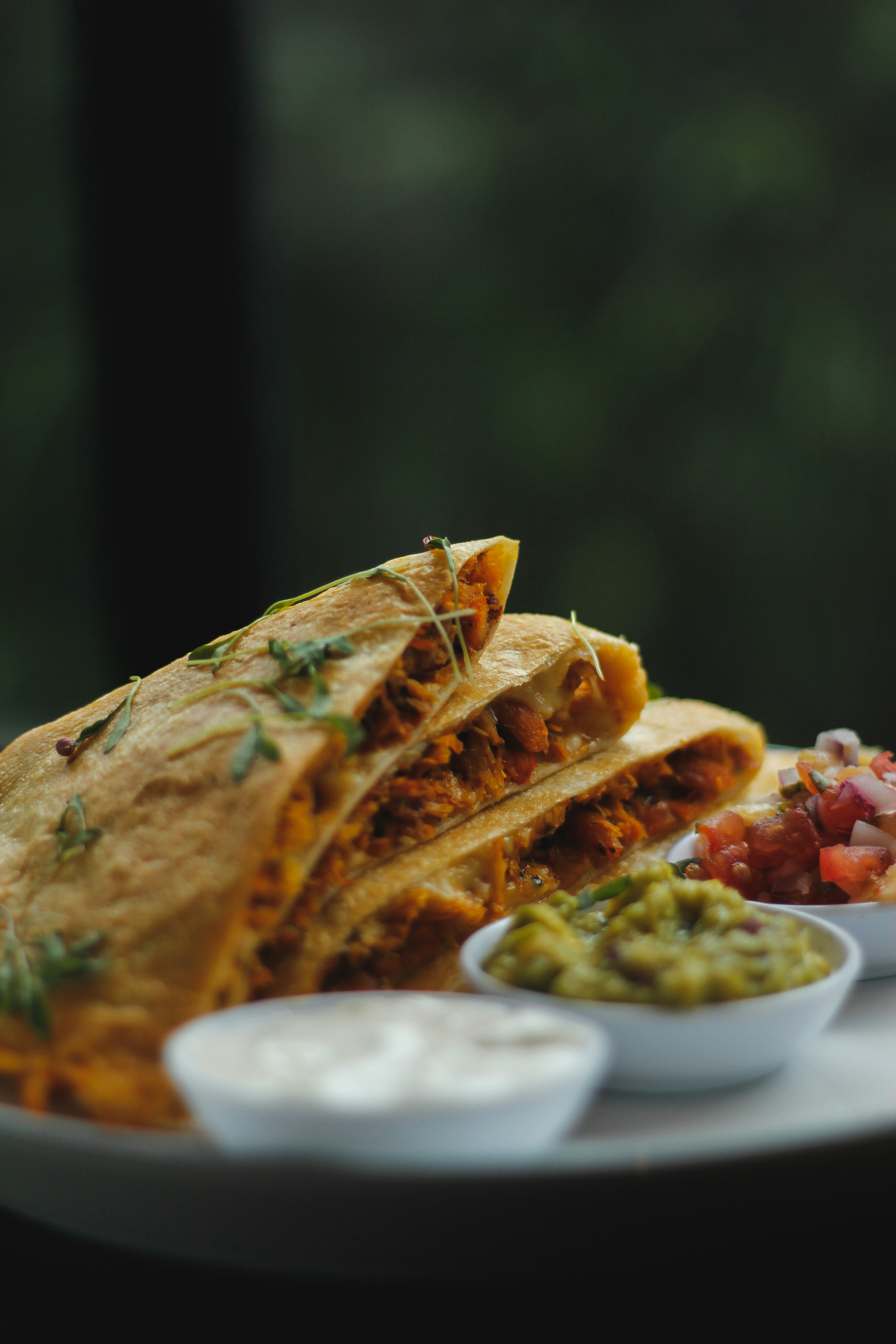 A stack of golden quesadillas garnished with herbs sits on a plate accompanied by bowls of guacamole, salsa, and sour cream