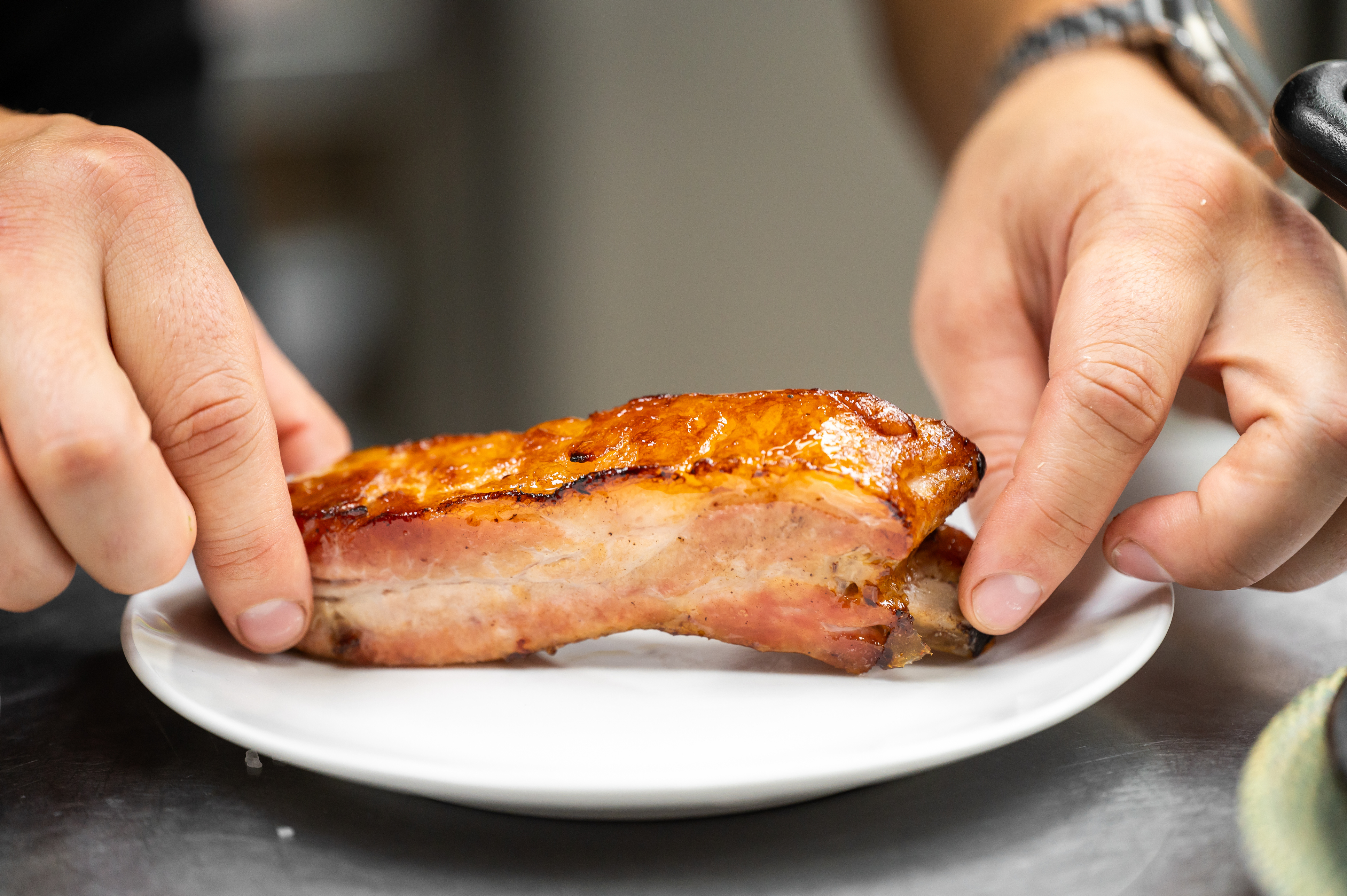 A pair of hands placing a piece of cooked pork belly onto a white ceramic plate