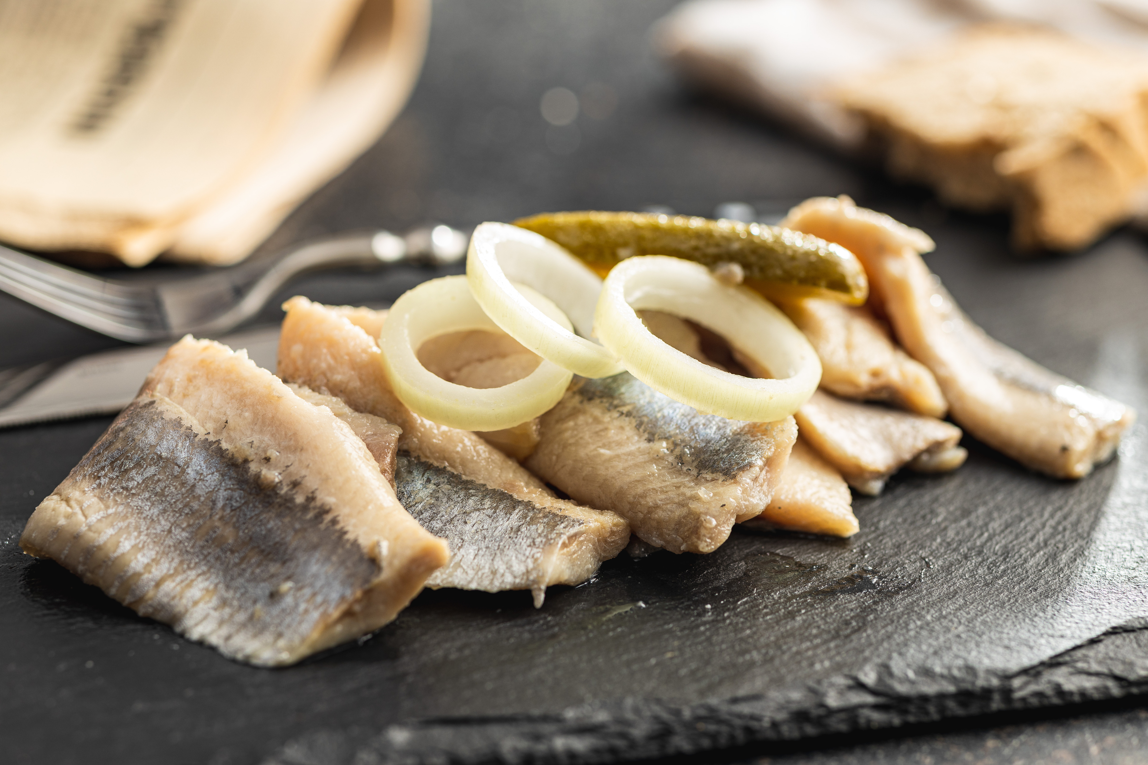 Sliced pickled herring fillets garnished with white onion rings and a gherkin on a dark slate board