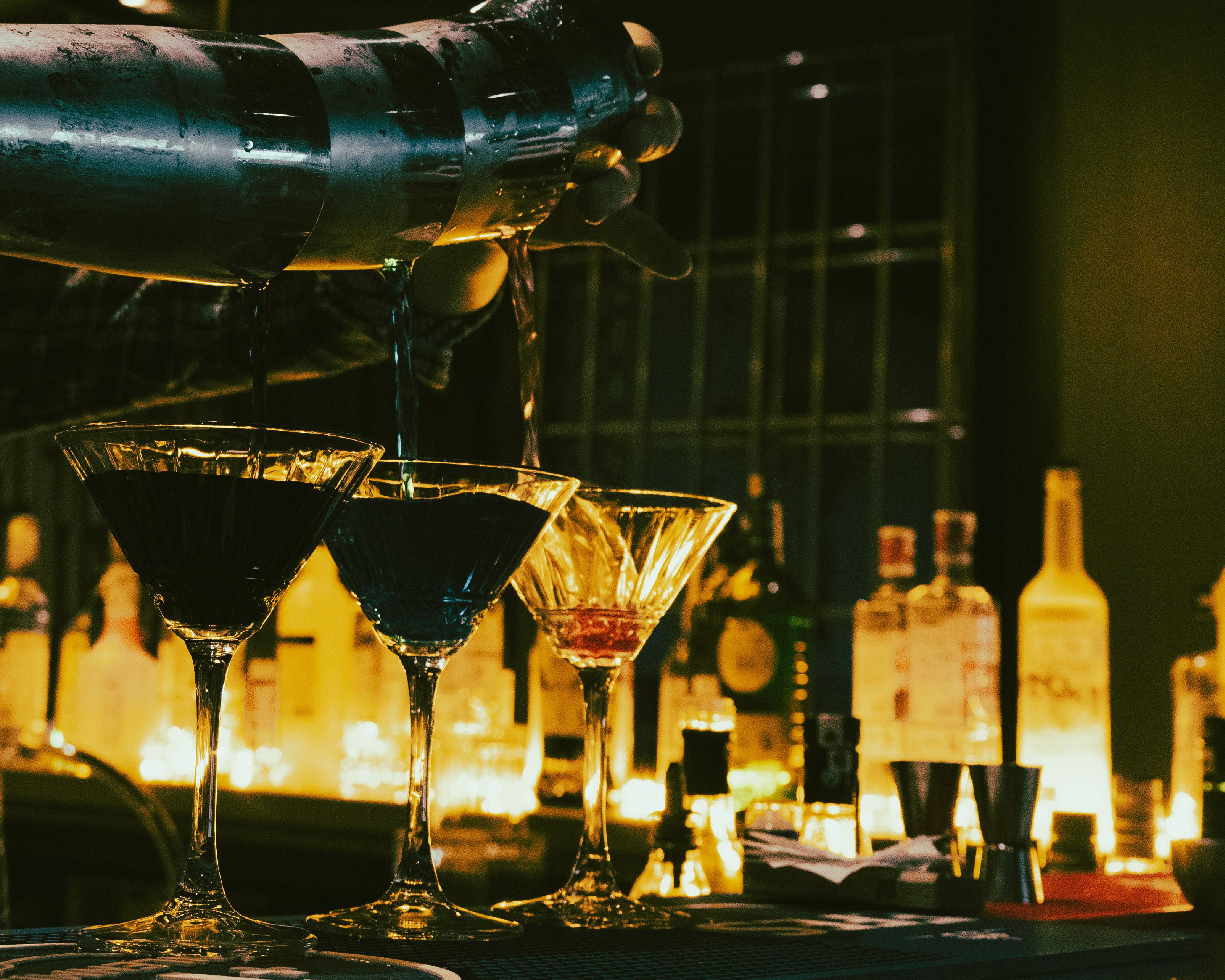 A bartender pouring a dark cocktail from a metal shaker into three martini glasses on a lit bar.