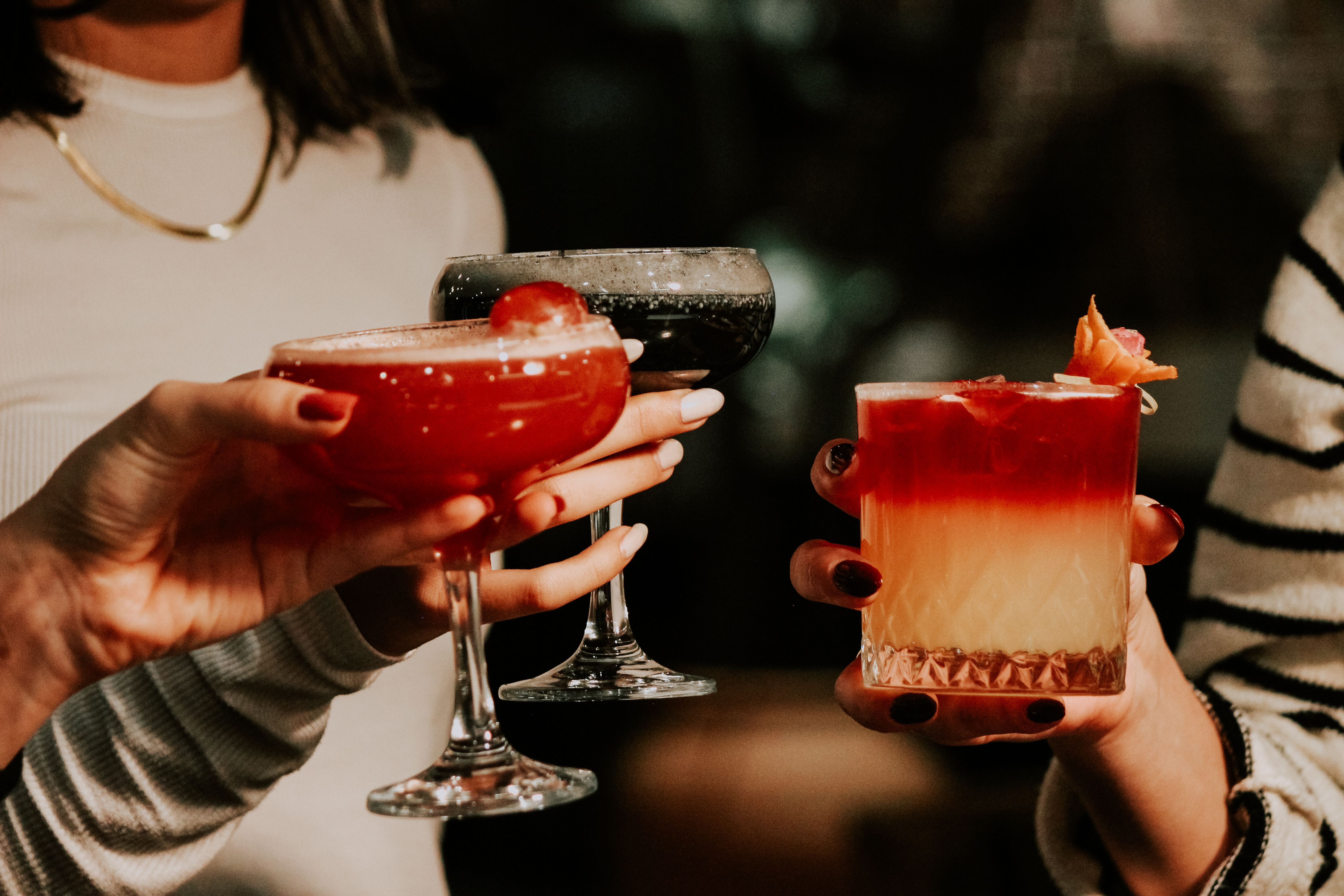 Three adults holding different colourful alcoholic cocktails in glasses